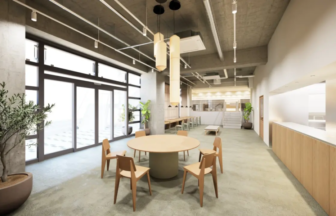 Open plan lounge/dining area with a round table and wooden chairs, large glass doors, and potted plants across a concrete ceiling space, soft lighting.]