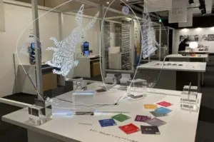 Interactive glass sculpture with two large text formed hands inside circular frames on a white display table in a gallery surrounded by colorful cards and clear blocksAcrylic exhibit two circular glass frames containing hand shaped text art over a white table with small blocks and color swatches in a museum settingTwo translucent rings hold hand shaped text art displayed on a table with acrylic blocks and colorful cards in a gallery environmentText based hand sculpture inside circular rims displayed on a white pedestal with educational cards in a modern museum spaceGallery exhibit featuring illuminated text made hand silhouettes within circular frames on a table with supporting blocks and swatches