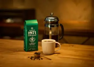 Green bag of Starbucks 1971 Dark Roast coffee on a wooden table beside a metal French press and a small white mug of coffee with coffee beans and a scoop in the foreground