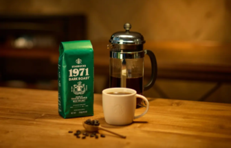 French press and a white mug of coffee on a wooden table with a green bag of Starbucks 1971 Dark Roast nearby.