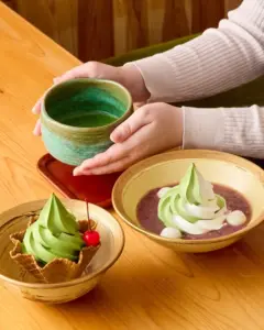 Hands cradle a green ceramic bowl over a wooden table surrounded by matcha desserts including green soft serve and red bean toppings