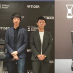 Three men in business attire pose beside a gray coffee grinder on a round table at a Tiger Hybrid Brew event