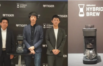 Three men in business attire pose beside a gray coffee grinder on a round table at a Tiger Hybrid Brew event.
