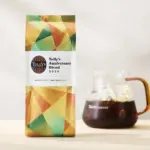 Bag of Tullys Coffee Anniversary Blend 2026 beside a glass carafe of dark brewed coffee on a light table