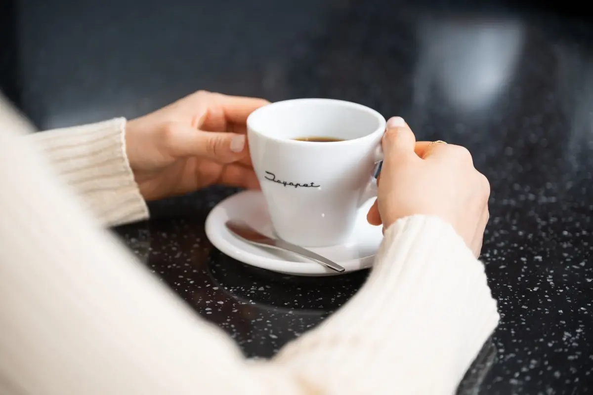 白いカップに入ったコーヒーを両手で持っている女性の手元が写っています。