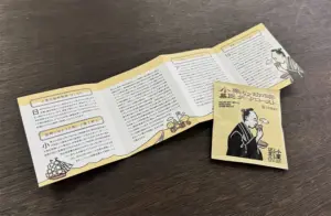 Accordion fold Japanese brochure spread on a dark surface with small illustrations and dense text panels plus a yellow sachet featuring a man in traditional dress