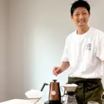 Smiling man in a white Tshirt and apron stands beside a pour over coffee setup with a copper kettle and filter dripper on the table
