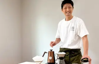 Smiling man in a white Tshirt and apron stands beside a pour over coffee setup with a copper kettle and filter dripper on the table