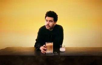 Man in a black jacket leans on a dark bar with a glass of iced coffee and a small cup of cream, against a yellow sunset backdrop.