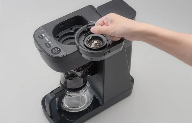 黒いコーヒーメーカーの蓋を片手が開けている様子