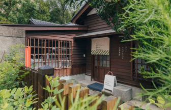 Wooden cafe building with a striped awning and Japanese signage, nestled among lush greenery outside the entrance.