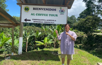Man standing outdoors next to a sign that says 'Bienvenidos al Coffee Tour,' pointing left in a banana garden setting.
