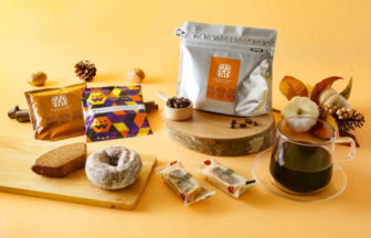 Autumn-themed coffee products and pastries arranged on a wooden board: a silver coffee bag, colorful Wrapped bars, a donut, bread slice, and a cup of black coffee against a yellow background.