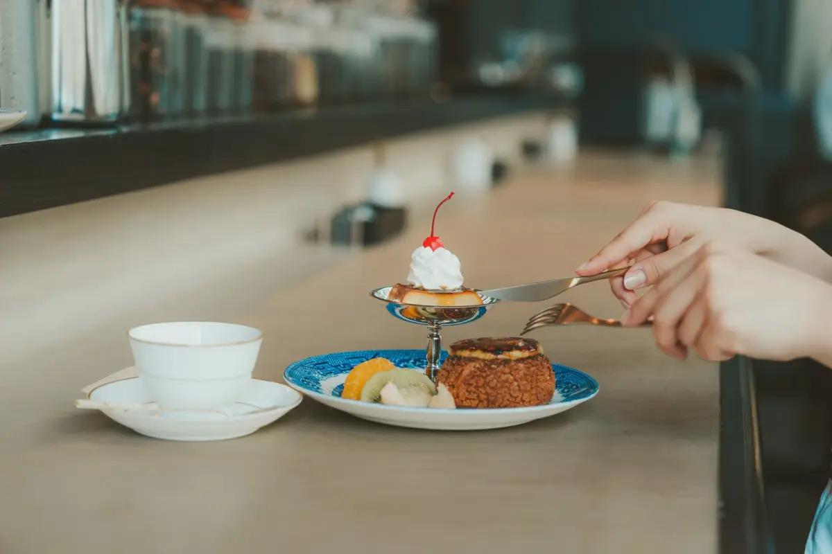 カフェで、女性がナイフとフォークを使って豪華なデザートをまさに食べようとしている様子です。プリンやシュークリーム、カットフルーツが美しく盛り付けられています。