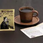 Steaming brown coffee cup on a saucer beside a yellow Japanese coffee sachet and an open brochure on a dark wooden table