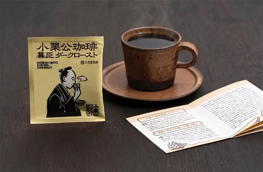 Steaming brown coffee cup on a saucer beside a yellow Japanese coffee sachet and an open brochure on a dark wooden table