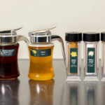 Two glass syrup decanters with metal pour spouts sit next to four labeled spice jars on a dark kitchen counter Ginger Sugar Chip Vanilla Sugar  Salt Cinnamon Powder