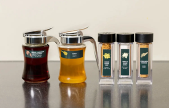 Two glass syrup decanters with metal pour spouts sit next to four labeled spice jars on a dark kitchen counter Ginger Sugar Chip Vanilla Sugar  Salt Cinnamon Powder