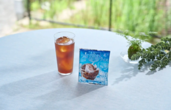 Tall glass of iced tea with ice on a white outdoor table, beside a blue product package and a small potted plant, with greenery in the blurred background.