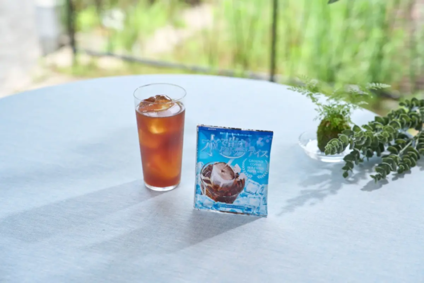 Tall glass of iced tea with ice on a white outdoor table beside a blue product package and a small potted plant with greenery in the blurred background
