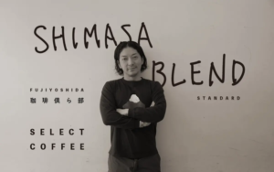 Man with arms crossed stands in front of a wall mural with large handwritten SHIMASA BLEND and SELECT COFFEE text giving a casual portrait look