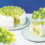 White frosted cake topped with green grapes sliced to reveal yellow cake layers and grape filling set against a blue backdrop with grape clusters nearby in view