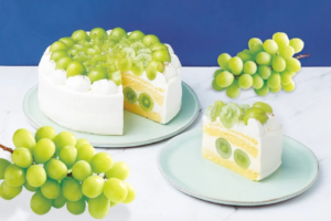 White frosted cake topped with green grapes sliced to reveal yellow cake layers and grape filling set against a blue backdrop with grape clusters nearby in view