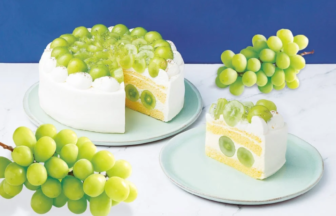 White frosted cake topped with green grapes, sliced to reveal yellow cake layers and grape filling, set against a blue backdrop with grape clusters nearby in view.