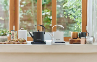 Two Russell Hobbs electric kettles (black and white) sit on portable stove tops on a bright kitchen counter with bread, jars, and plants nearby by a sunlit window.