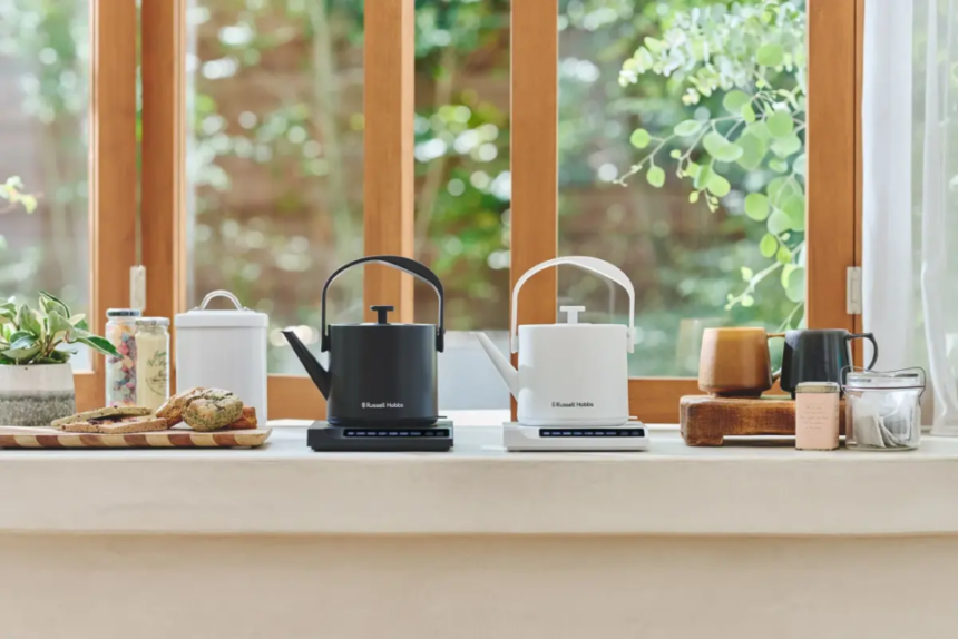 Two Russell Hobbs electric kettles black and white sit on portable stove tops on a bright kitchen counter with bread jars and plants nearby by a sunlit window