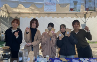 Five people stand behind an outdoor market booth, smiling and holding brown paper bags with circular labels; coffee equipment sits on the table in front of them.