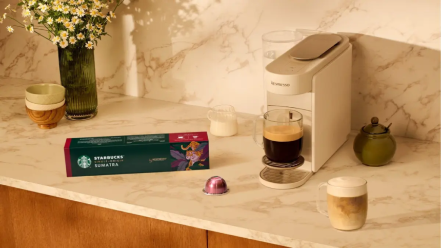 Marble kitchen counter with a Nespresso machine a glass mug of coffee a Starbucks Sumatra box and a pink coffee capsule beside a vase of daisies