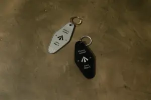 Two metal key fobs on a dusty floor one gray and one black each with a paperairplane icon and the messages Cheers buddy and Love Nige×