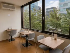 93890a911cdf8fc0d92f63e5cfe735f4 | コーヒーステーション Café seating area with three wooden tables and light gray chairs by a large window overlooking green trees and modern buildings outside