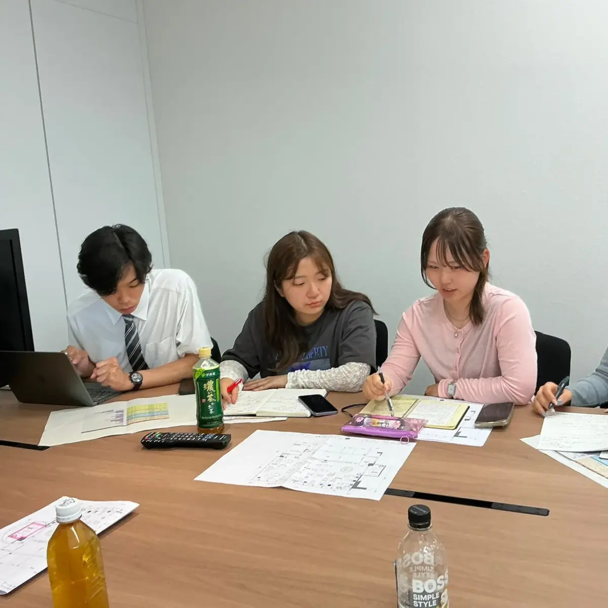 若者3人が会議室のような空間でノートやパソコン、資料を広げて話し合いや作業をしている様子