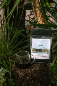 990cf33f6459367772f5f84a8727ab4a | コーヒーステーション Bag of coffee labeled Japan Tokunosima CWS beside a glass carafe of dark brew on a tree stump among tropical plants