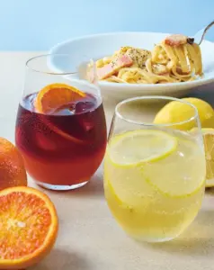 Two drinks and a pasta dish on a light table red sangria with an orange slice and a lemon soda with lemon wheels surrounded by citrus fruits