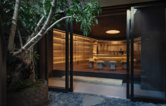 Entrance to a stylish interior bar seen through open glass doors, with a circular, warmly lit counter and beige stools against curved wooden shelves in back.