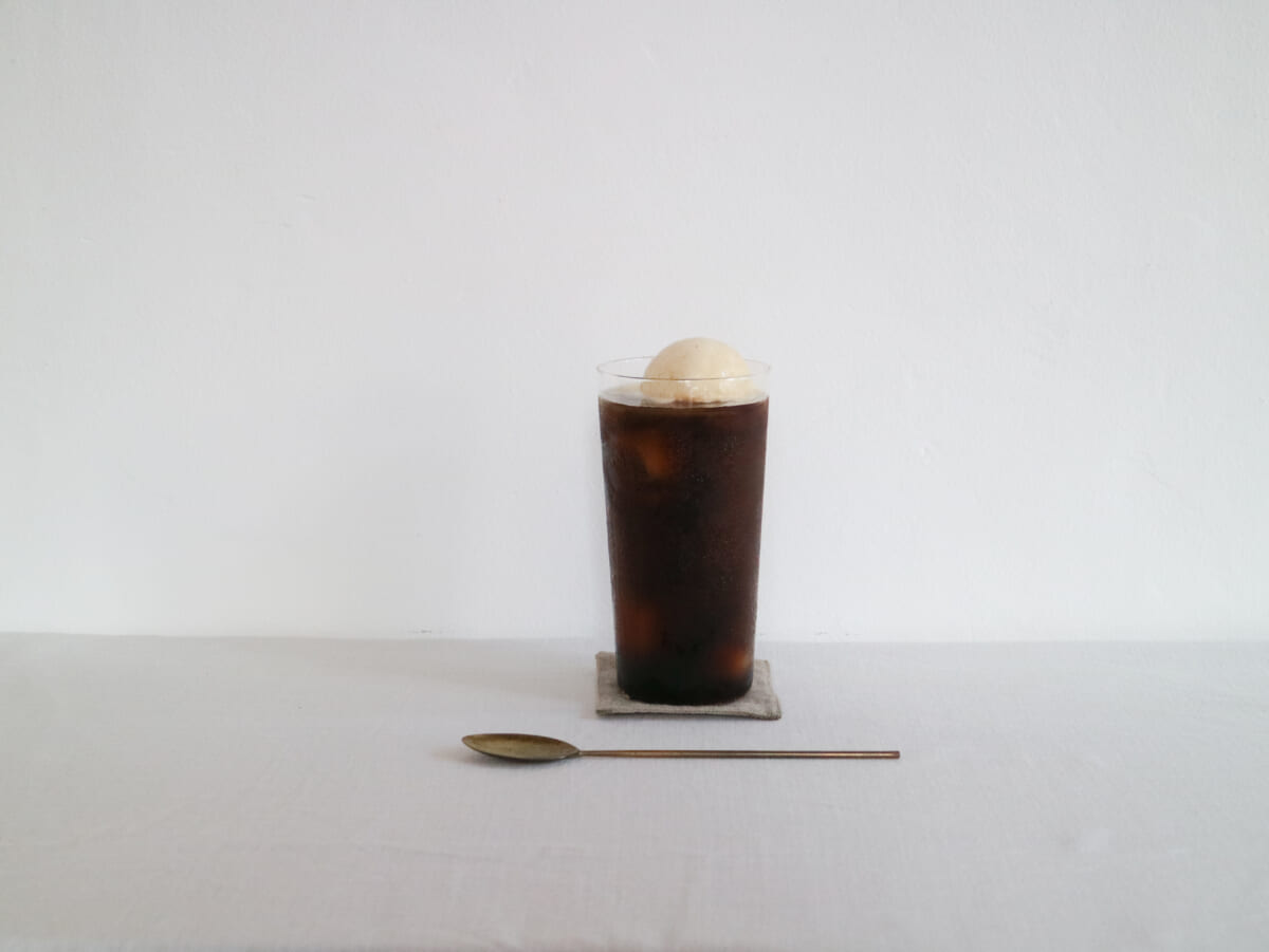 Tall glass of iced coffee with a scoop of vanilla ice cream on top placed on a small coaster against a white background with a spoon in the foreground