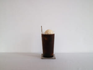 Tall glass of iced coffee with a scoop of vanilla ice cream on top placed on a small coaster against a white wall