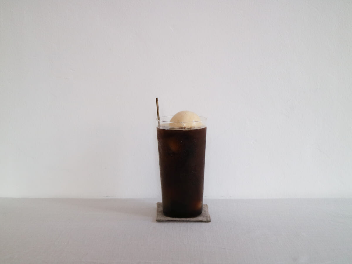 Tall glass of iced coffee with a scoop of vanilla ice cream on top placed on a small coaster against a white wall