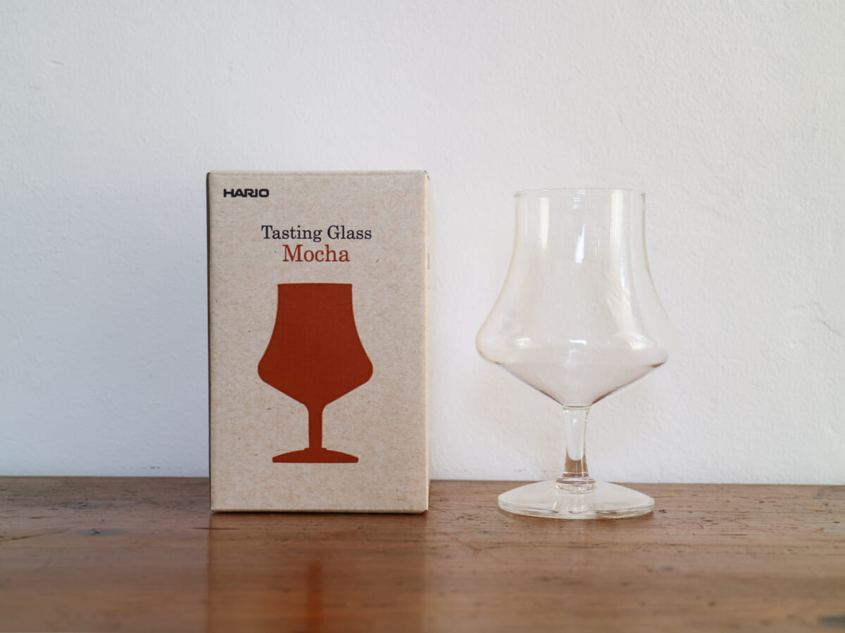 IMG4227 | コーヒーステーション Box labeled HARIO Tasting Glass Mocha next to a clear stemmed glass on a wooden table against a pale wall