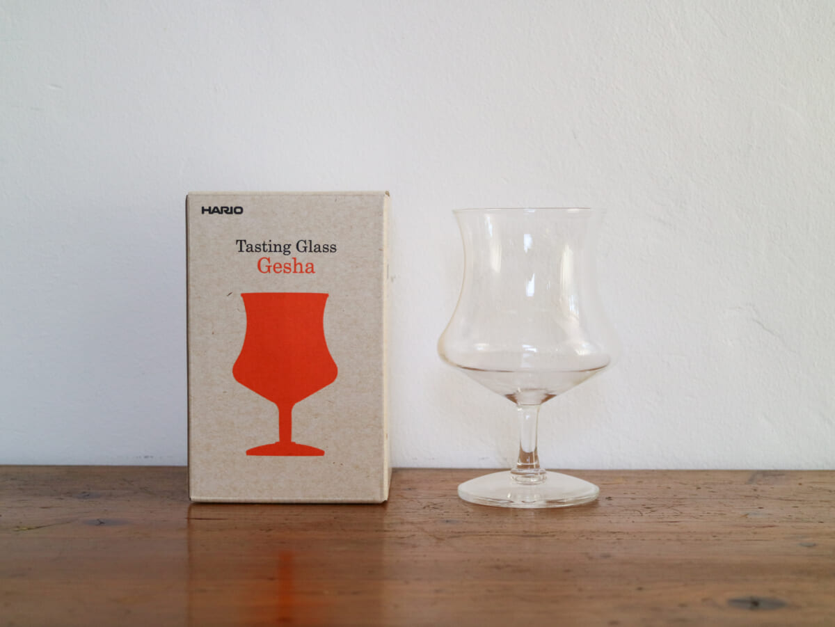 IMG4228 | コーヒーステーション Box labeled'Tasting Glass Gesha' beside a clear glass tasting goblet on a wooden table surface.