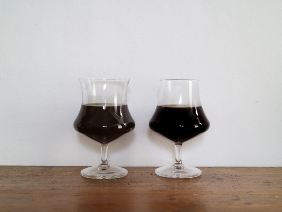 IMG4393 | コーヒーステーション Two snifter glasses of dark beer on a wooden table against a white wall side by side