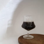 A snifter glass with dark stout on a wooden round table against a plain white wall