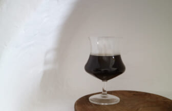 A snifter glass with dark stout on a wooden round table against a plain white wall