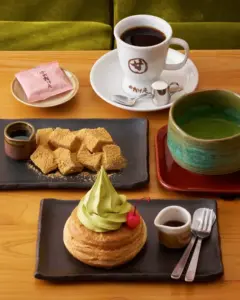 Cafe table with matcha soft serve on pastry topped with a cherry a plate of kinako dusted squares a cup of coffee and a green tea bowl on a red tray