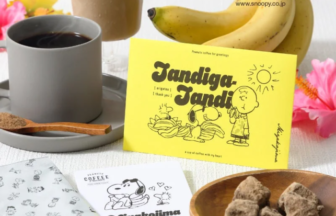 Coffee scene with a gray mug of black coffee, a wooden spoon, a glass of iced coffee, yellow Peanuts card, bananas, and a bowl of brown sugar cubes on a white table.