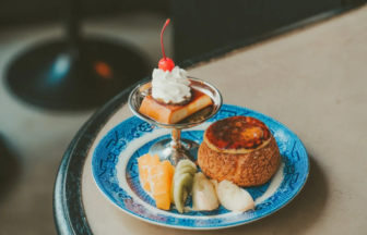 Blue patterned plate with a fruit assortment orange kiwi banana and a mini crème brûlée topped with whipped cream and a cherry in a cocktail glass on the side