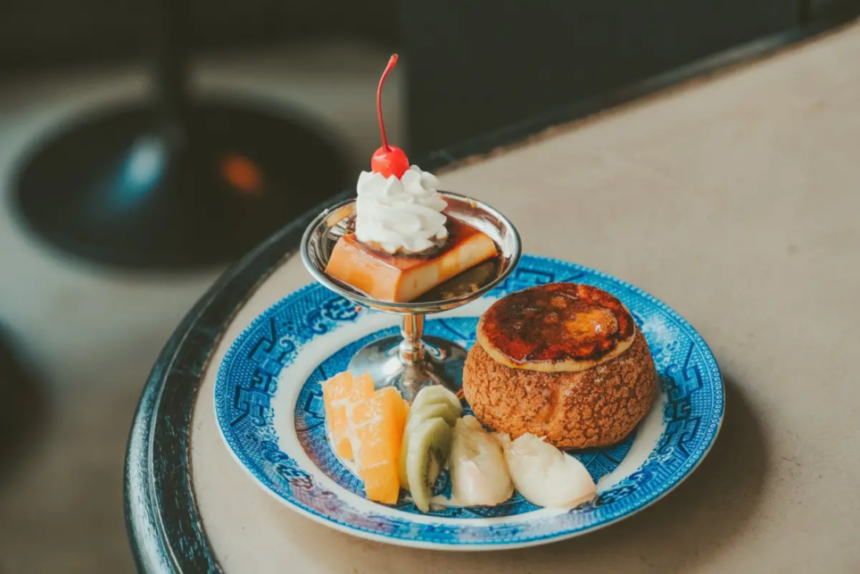Blue patterned plate with a fruit assortment orange kiwi banana and a mini crème brûlée topped with whipped cream and a cherry in a cocktail glass on the side